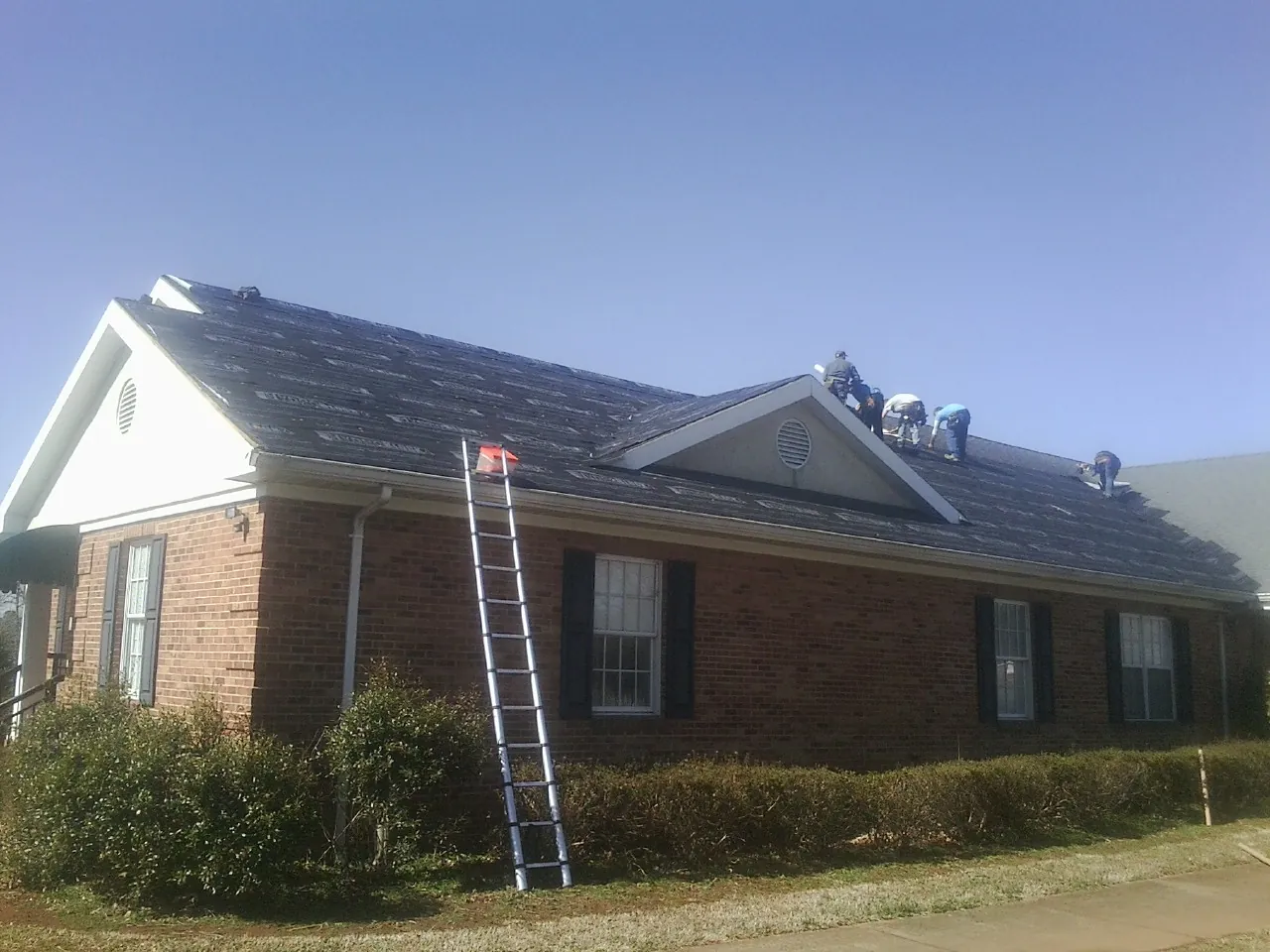Crew tearing shingles off a brick ranch roof with a ladder set — Emergency Roof Repair work by our crew in Saint Johns